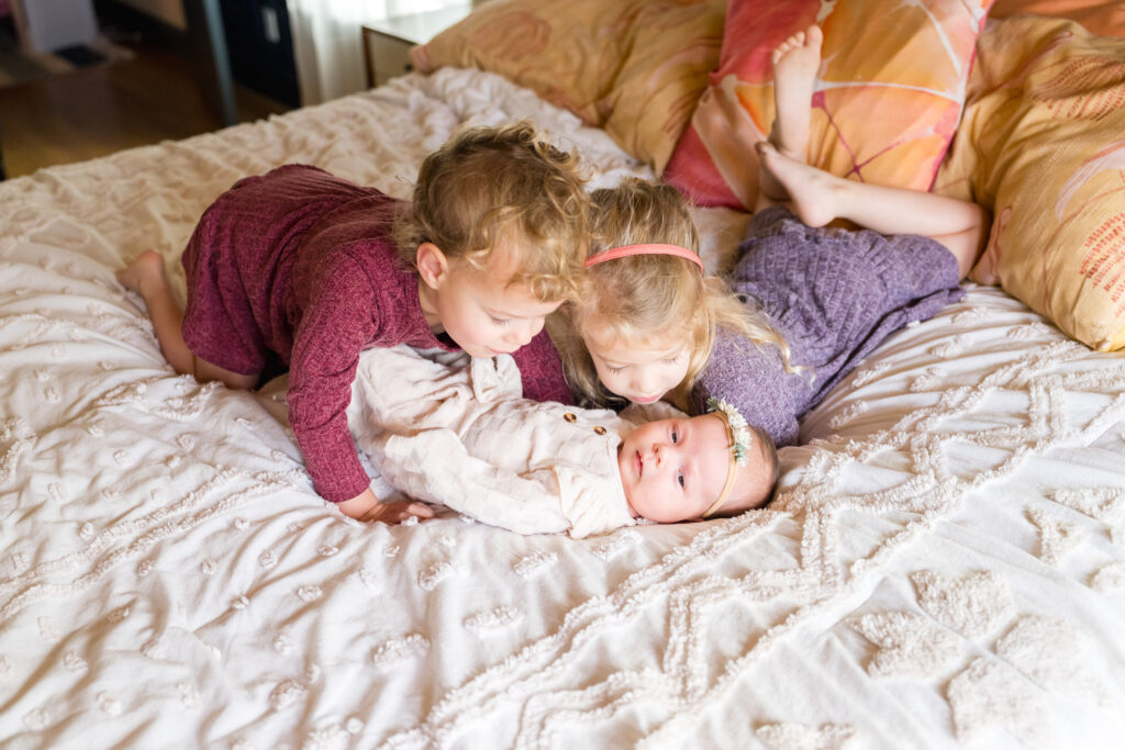 Three sisters look lovingly at their newborn sibling during a newborn photoshoot.