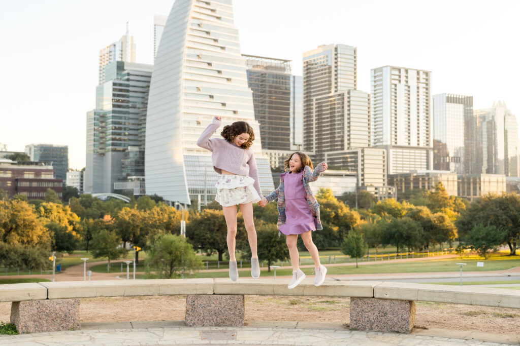 The beautiful Austin skyline in the background as two sisters jump in the air, holding hands. 