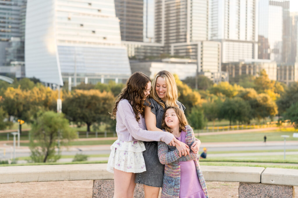 A mom and her two daughters hugging each other and smiling, with the Austin skyline behind them.