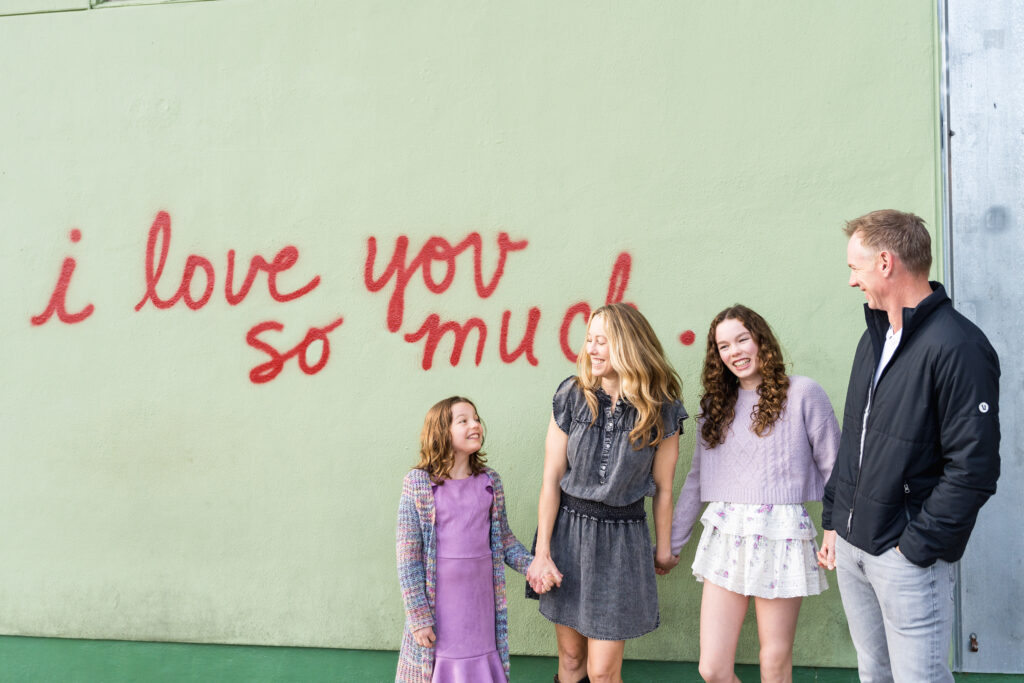 A family holding hands and laughing during their family photo session with a colorful wall in Austin behind them.