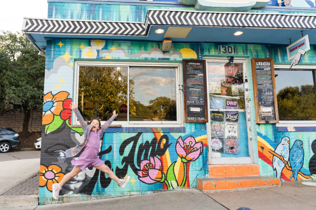 A pre-teen aged girl jumping happily in the air in front of a colorful downtown cafe in Austin, Texas.