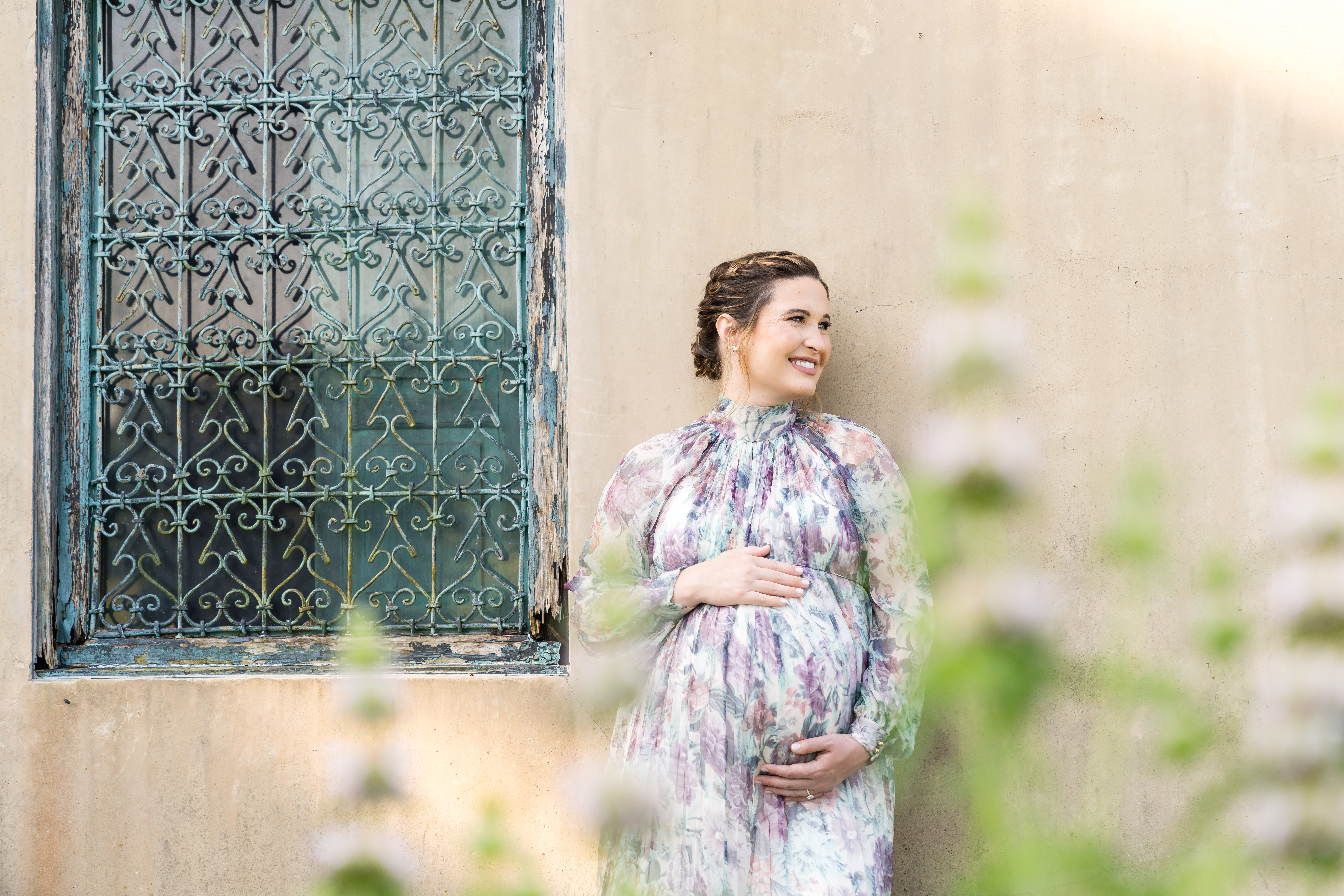 pregnant mom wearing a floral dress, while wrapping her arms around her belly and leaning up against a building in Austin, TX