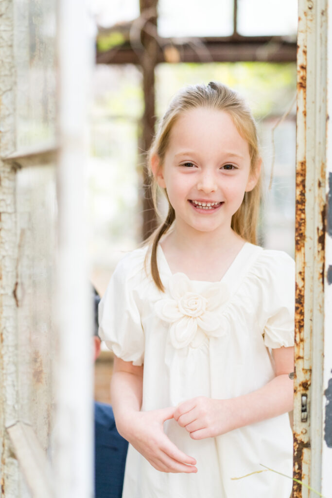 A young girl wearing a cream dress, peeking through an open door during her family photos in Austin.