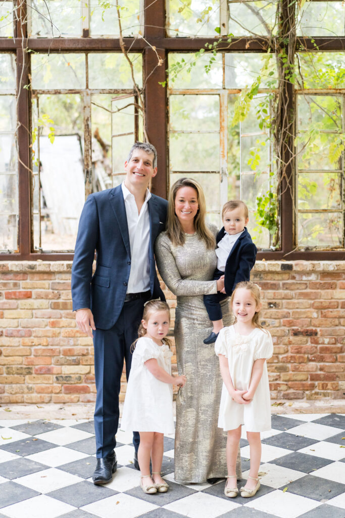 A mom and dad with their 3 children, having their family photos in a greenhouse with walls made of bricks and windows, in Austin, Texas.