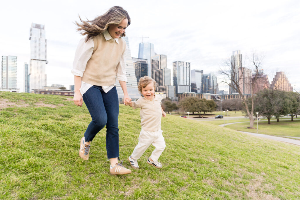 A mom running down a grassy hill with her son during their family photo session at Zilker Park.