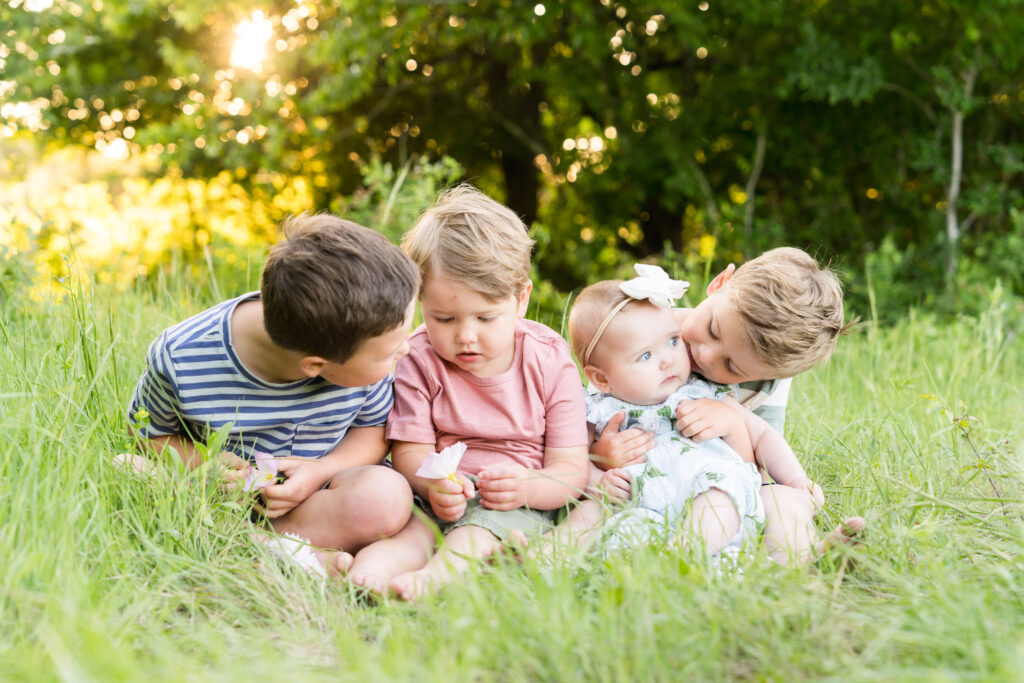 A 6 month old girl and her 3 older brothers sitting in the grass during her Baby's First Year photo session.
